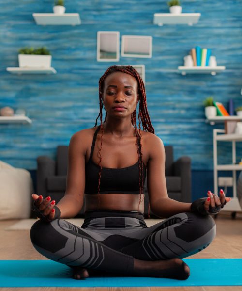 Black woman training body and mind meditating in lotus pose keeping eyes closed, sitting on yoga mat in home living room for calm, healthy harmony lifestyle dressed in sportwear.