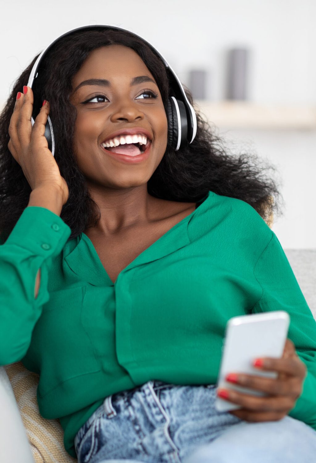 Positive attractive millennial african american woman in casual with wireless headset and cellphone sitting on couch at home, listening to music, looking at copy space and smiling