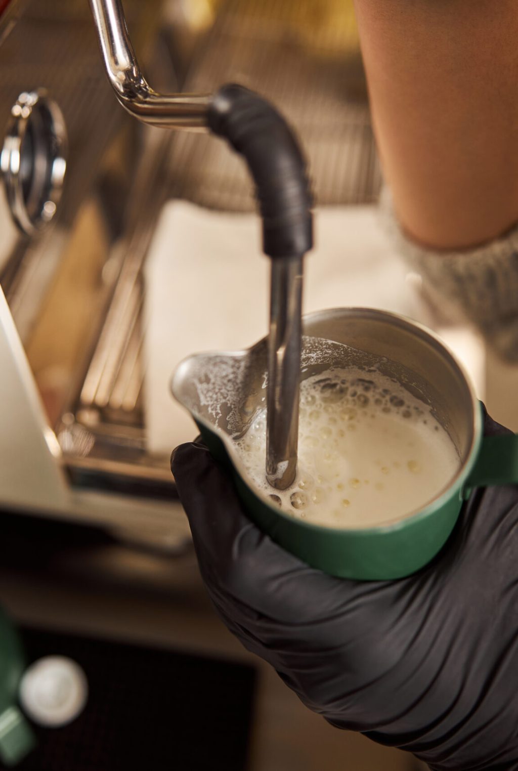 Barista in rubber gloves holding a pitcher with milk and using a steam wand for stirring it